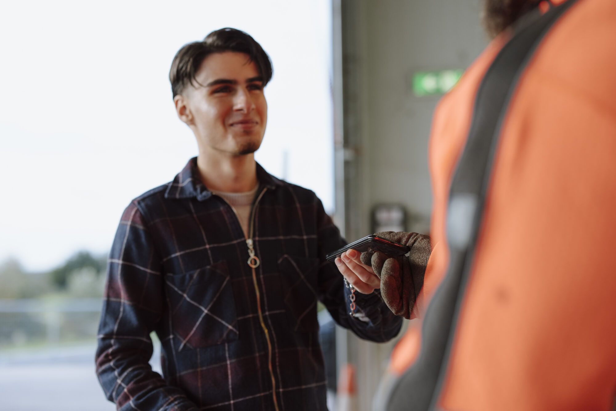 Ein junger Mann mit braunen Haaren und einer blau-lila Weste übergibt einem ASZ/WSZ Mitarbeiter, der nur mit einer Jacke abgebildet ist, ein Handy.