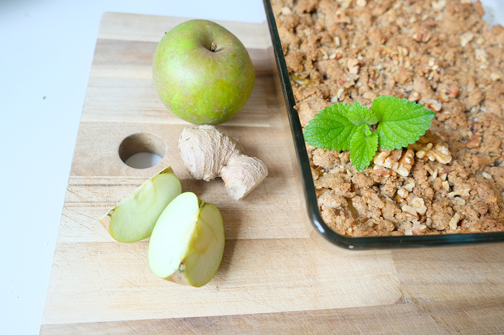 Auf einem Holzbrett liegen ein grüner Apfel, ein Stück Ingwer und zwei Apfelspalten. Daneben ist eine Glasauflaufform mit Apfel-Crumble, garniert mit einem Zweig Zitronenmelisse