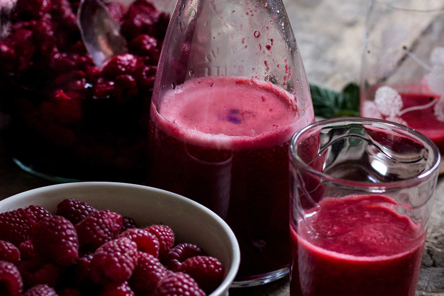 Eine Schüssel Himbeeren verschiedene Gläser gefüllt mir leuchted rotem Himbeersaft.